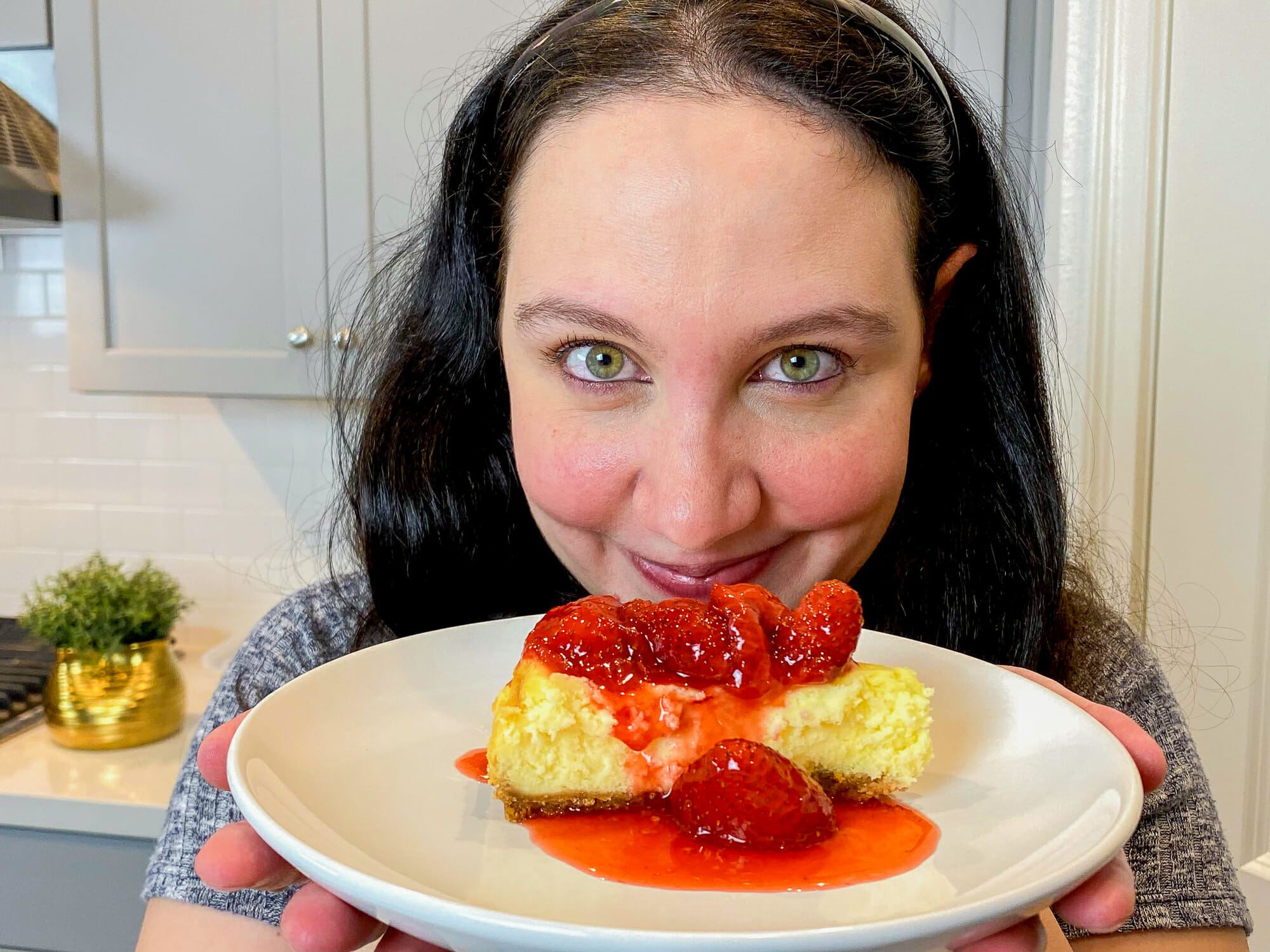 Foto da Renata segurando a fatia de cheesecake da Cheesecake Factory em frente à câmera e sorrindo. A cheesecake está coberta por morangos e calda de morango.