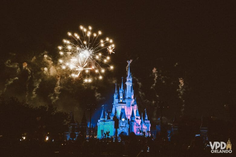 Foto do show de fogos do Magic Kingdom, com o castelo iluminado em azul e rosa, o céu escuro e os fogos estourando. 