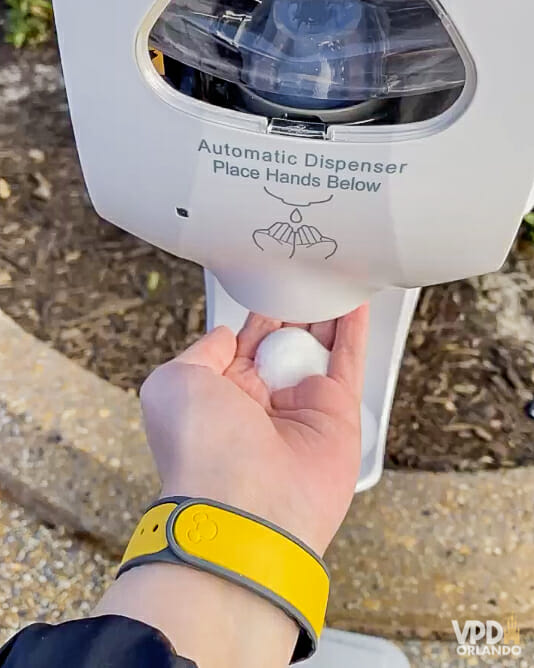 Foto de uma mão com uma MagicBand amarela no pulso pegando sabão em um dispenser automático na estação de higienização 