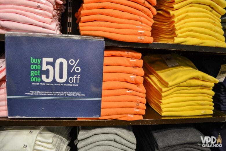 Foto de pilhas de camisetas de várias cores à venda em um outlet, com uma placa indicando 50% de desconto 