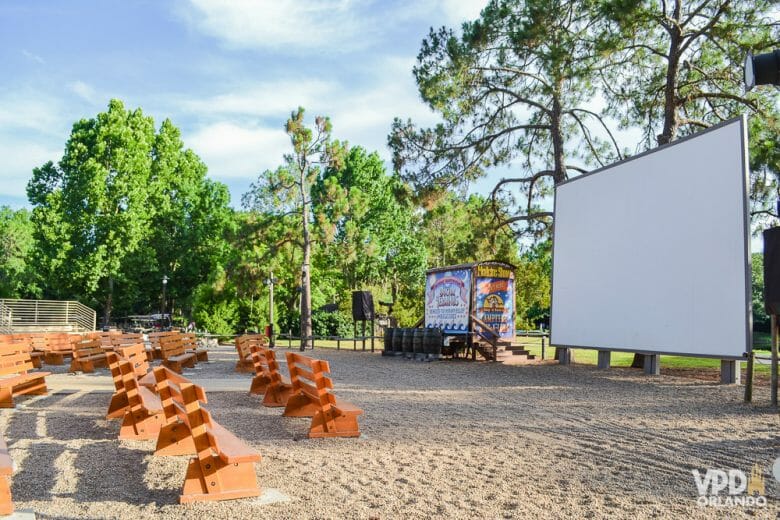 Foto do cinema a céu aberto no hotel Fort Wilderness. Há bancos para os visitantes sentarem e um telão em branco sobre a areia, com árvores em volta. 