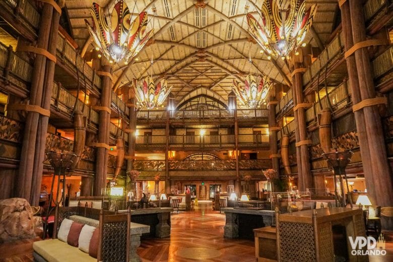 A imagem mostra o lobby do Animal Kingdom Lodge, um dos hotéis de luxo da Disney. A decoração é toda em madeira, com pilares, teto alto e lustres coloridos.