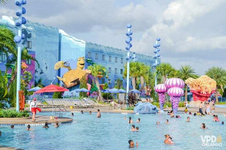 A imagem mostra visitantes na piscina do hotel da Disney, o Art of Animation. A decoração é colorida, o prédio ao fundo é azul e a tartaruga do filme Procurando Nemo é parte da decoração. 