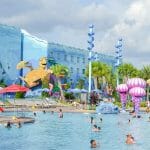 A imagem mostra visitantes na piscina do hotel da Disney, o Art of Animation. A decoração é colorida, o prédio ao fundo é azul e a tartaruga do filme Procurando Nemo é parte da decoração.