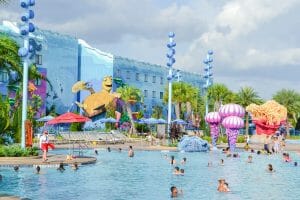 A imagem mostra visitantes na piscina do hotel da Disney, o Art of Animation. A decoração é colorida, o prédio ao fundo é azul e a tartaruga do filme Procurando Nemo é parte da decoração.