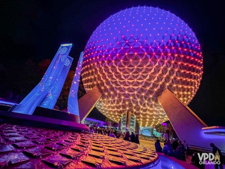 Foto da bola do Epcot iluminada em rosa e roxo à noite, com a fonte em frente.