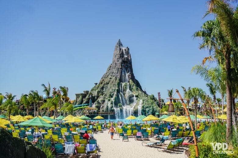 Imagem do vulcão que é símbolo do Volcano Bay, com as cadeiras de praia e guarda-sóis dos visitantes ao redor e o céu azul ao fundo. 