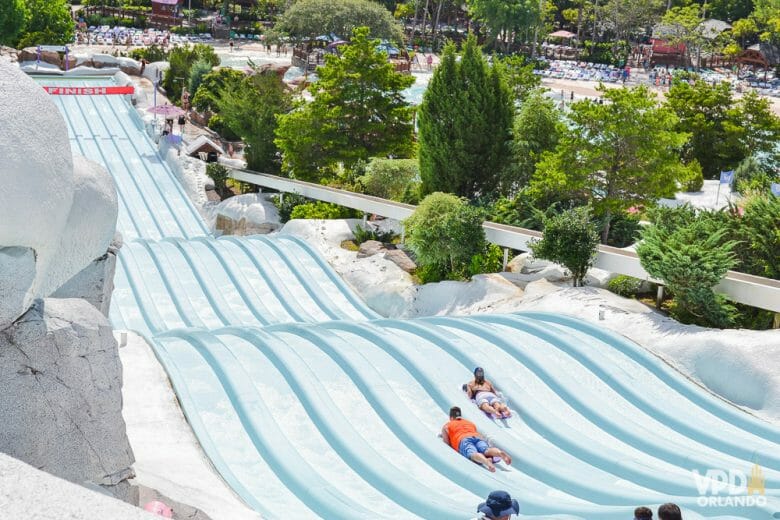Imagem do tobogã do Blizzard Beach, um dos  parques aquático da Disney. Os visitantes estão descendo o tobogã azul-claro, que é longo e tem decoração de neve ao redor. 