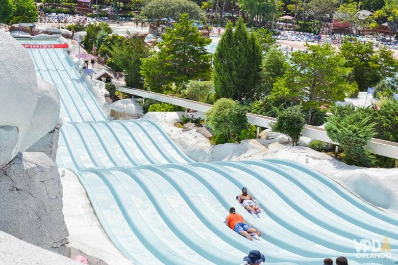 Imagem do tobogã do Blizzard Beach, um dos  parques aquático da Disney. Os visitantes estão descendo o tobogã azul-claro, que é longo e tem decoração de neve ao redor. 