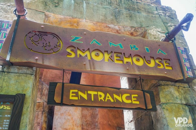 Opção gostosa de restaurante no Busch Gardens. Foto da placa na entrada do restaurante Zambia Smokehouse, em letras verde e amarelas sobre um fundo marrom 