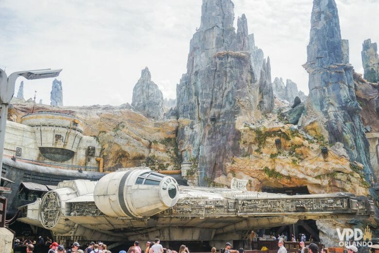 A Star Wars: Galaxy's Edge fica no Hollywood Studios. oto da Millenium Falcon com alguns visitantes ao redor, e parte da ambientação da Galaxy's Edge do Hollywood Studios visível ao fundo. 