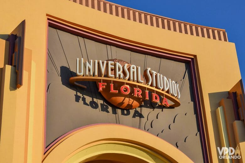 Foto do portal do parque Universal Studios Florida, que mostra a placa com o nome do parque escrito em branco e laranja. O portal é pintado em bege e laranja-claro e o céu ao fundo está bem azul.
