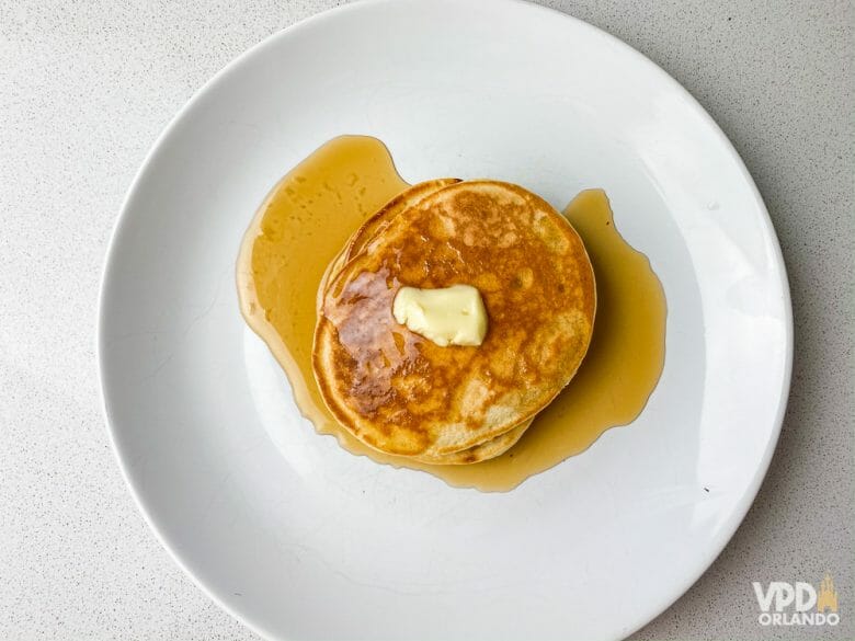 Você também vai precisar de maple syrup, manteiga, nutella ou o que mais quiser usar pra servir a sua panqueca! Foto de um prato branco com duas panquecas americanas, com um pouco de manteiga em cima e xarope de maple embaixo. 