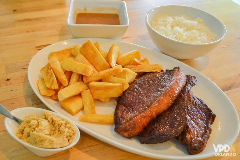 O Ana's Kitchen tem porções fartas e comidinha bem brasileira! É para não ter saudades de casa e ao mesmo  tempo não precisar gastar demais! Foto do prato no Ana's Kitchen, com dois pedaços de picanha ao lado de batatas fritas, arroz, farofa e molho 