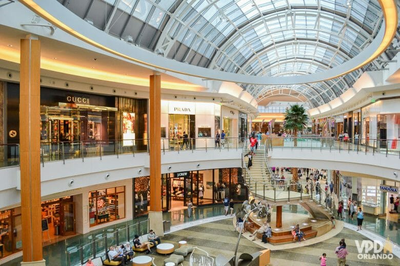 Imagem de um shopping. O centro do shopping tem visitantes caminhando e é possível ver as lojas de dois andares, bem como a abóbada transparente que permite a iluminação. 