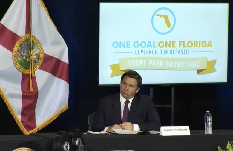 Foto do governador da Flórida, DeSantis, na reunião One Goal One Florida, que definiu a capacidade permitida em parques temáticos. Ele está sentado em uma mesa, com um telão atrás. 