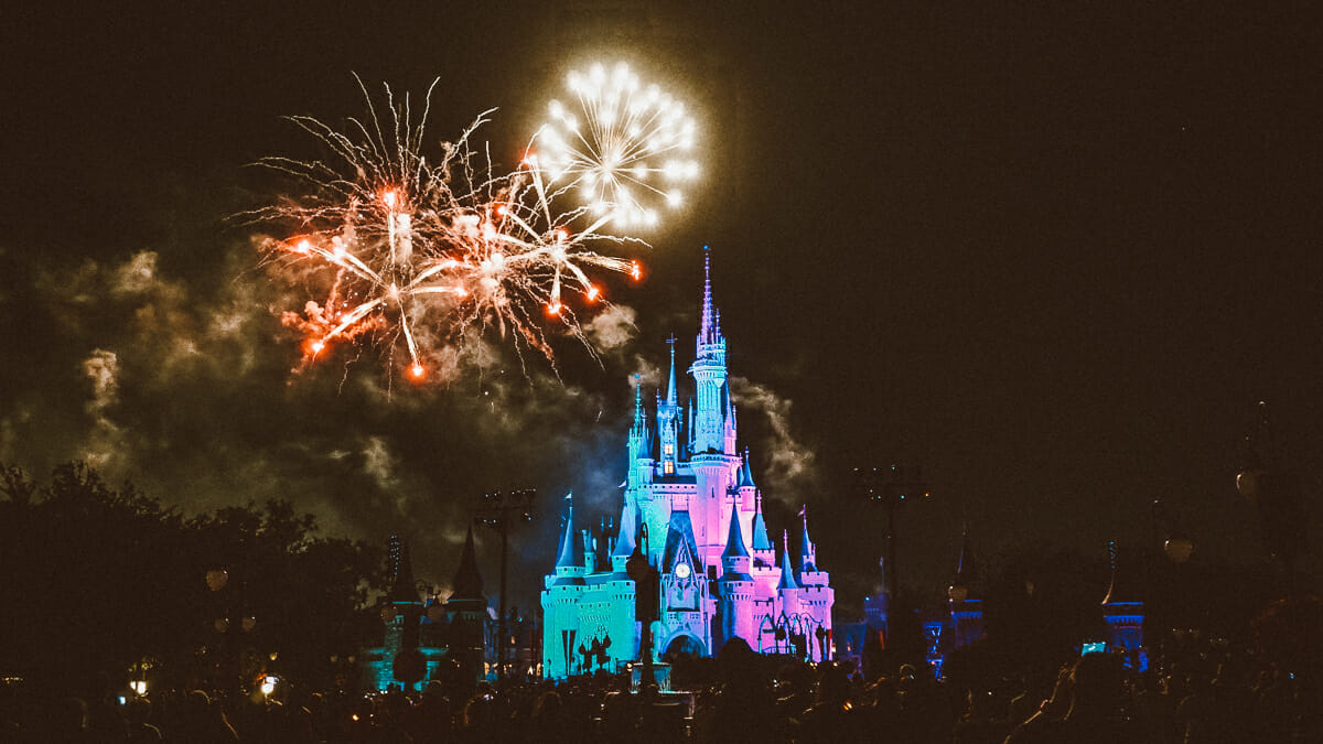 Imagem do Castelo da Cinderela no Magic Kingdom à noite, iluminado pelos fogos ao fundo e pelas luzes coloridas.