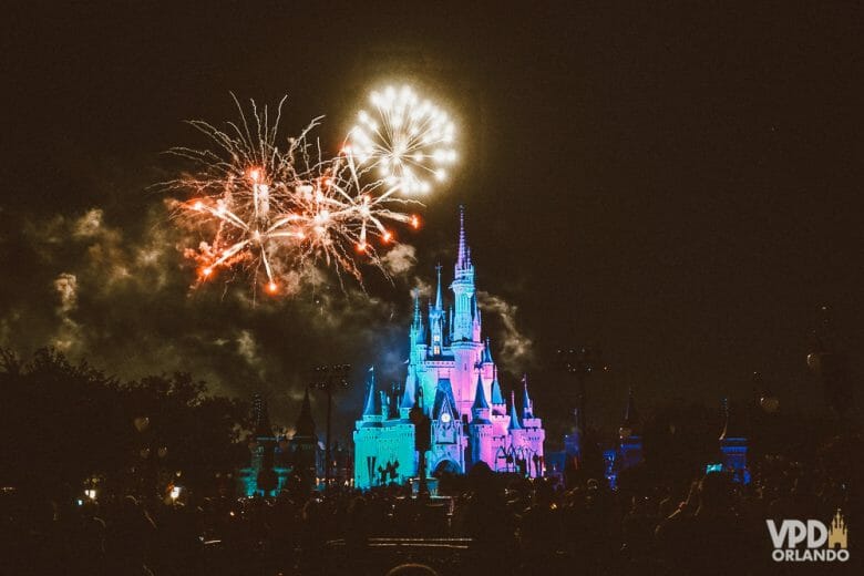 Foto do Magic Kingdom durante o show de Fogos Happily Ever After. O castelo da Cinderela brilha com luz azul e cor-de-rosa e os visitantes estão na sombra. Um dos maiores erros é perder isso e ficar filmando no celular! 