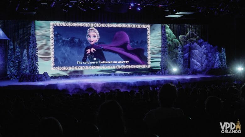 Imagem do palco durante o show de Frozen apresentado no Hollywood Studios, com a rainha Elsa no telão e a legenda para que os visitantes cantem junto. 