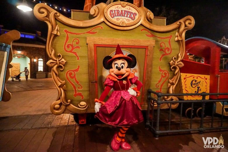 Imagem da Minnie posando em frente a uma carruagem na festa do Magic Kingdom. Ela está fantasiada de bruxa, com uma roupa vermelha e chapéu pontudo. 