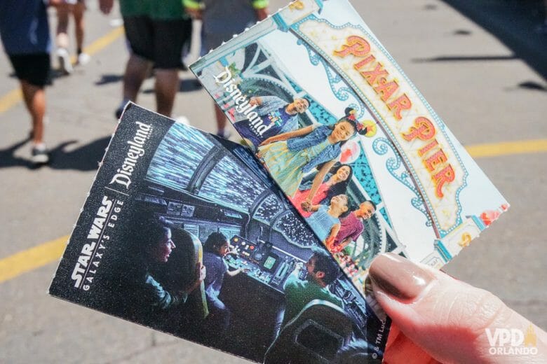 Imagem de uma mão segurando os ingressos em papel da Disneyland Califórnia, ilustrados com fotos das áreas do parque (Pixar Pier e Star Wars Galaxy's Edge). 