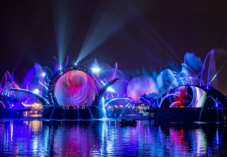 Foto de divulgação das luzes do HarmonioUS, o novo show noturno do Epcot, sobre o lago e com a Spaceship Earth ao fundo, em tons de azul, roxo e rosa 