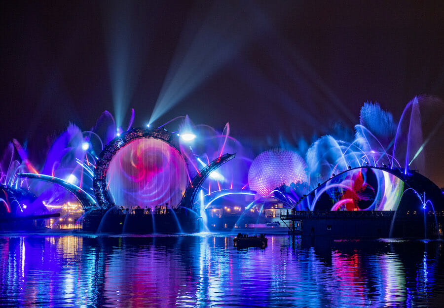 Foto de divulgação das luzes do HarmonioUS, o novo show noturno do Epcot, sobre o lago e com a Spaceship Earth ao fundo, em tons de azul, roxo e rosa