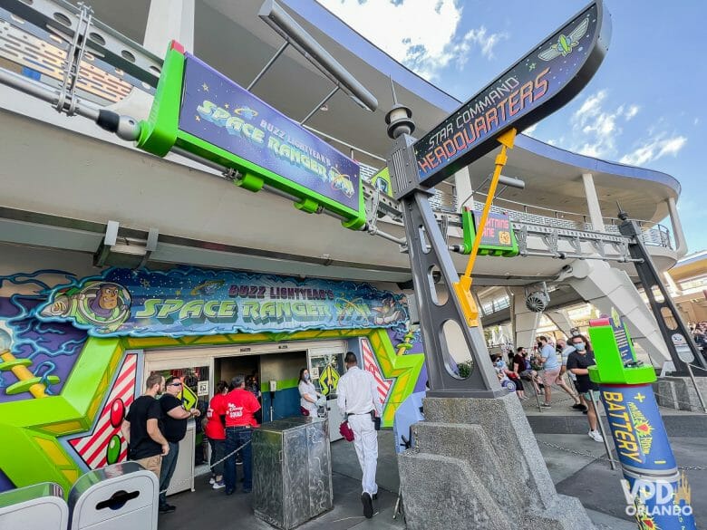 Foto wide-angle da fachada do Buzz Lightyear Space Ranger Spin. Há várias pessoas na entrada, e a fachada possui ilustrações em verde e azul. Essa atração do Magic Kingdom faz parte do Genie+.