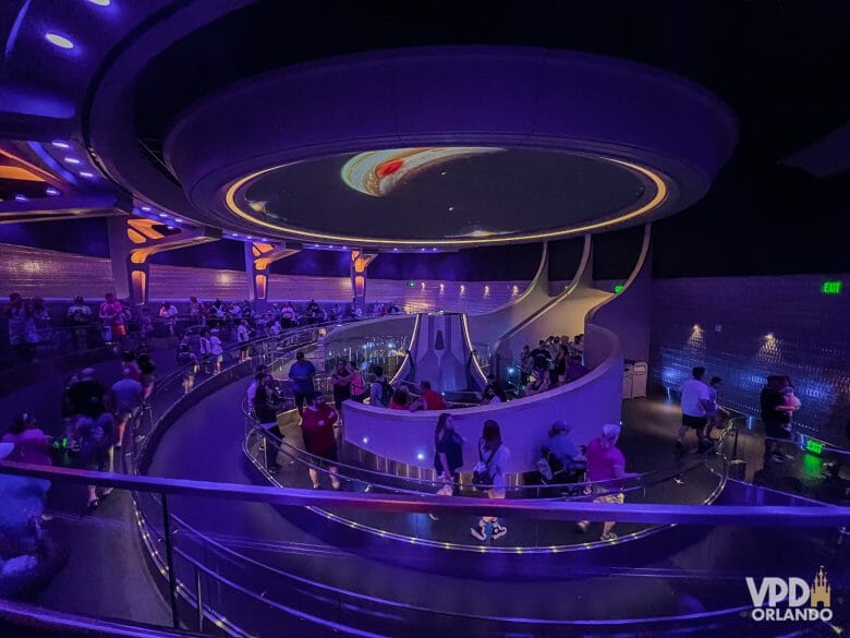 Foto de dentro do pavilhão da nova-montanha-russa. Há pessoas na fila, ele está iluminado em tons de roxo e há um planetário bem no meio.