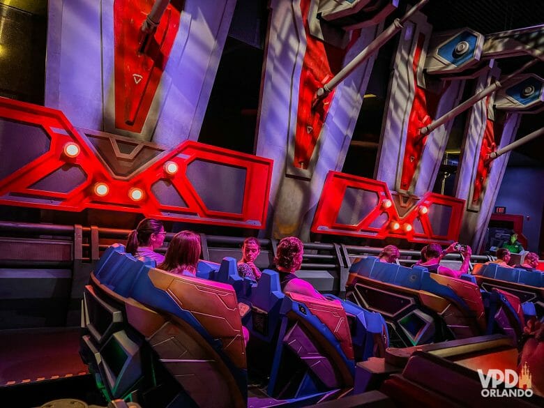 Foto da área de embarque da montanha-russa Guardians of the Galaxy: Cosmic Rewind. Os carrinhos são azuis e amarelos e há pessoas sentadas. As paredes possuem detalhes em vermelho.