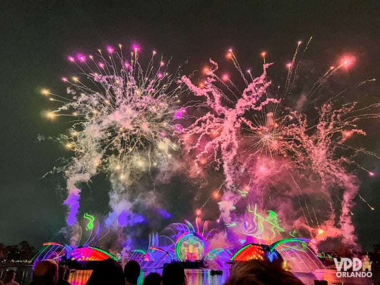 Foto do show de fogos Harmonious: há projeções coloridas, em vermelho, azul e verde nas estruturas no lago, e fogos coloridos estão estourando.