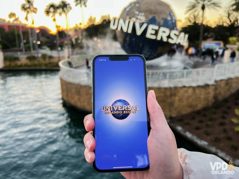 Foto de um celular com a tela inicial do aplicativo da Universal. Ao fundo, a bola com o logo da Universal está visível.