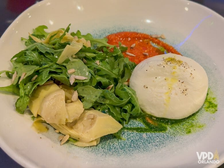 Prato com burrata, um molho vermelho, rúcula e alcachofras.