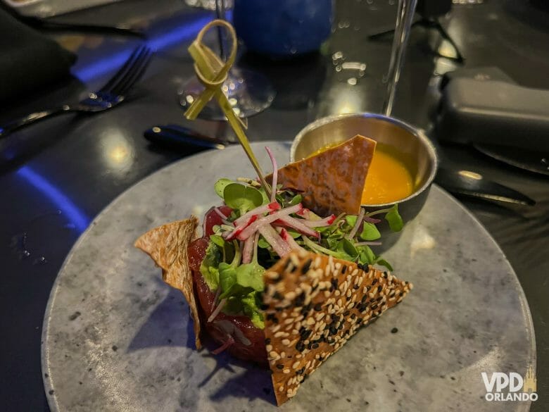 Entrada do restaurante, um tartar de atum com biscoitinhos com gergelim. Ao fundo há um potinho com uma calda amarela de manga.