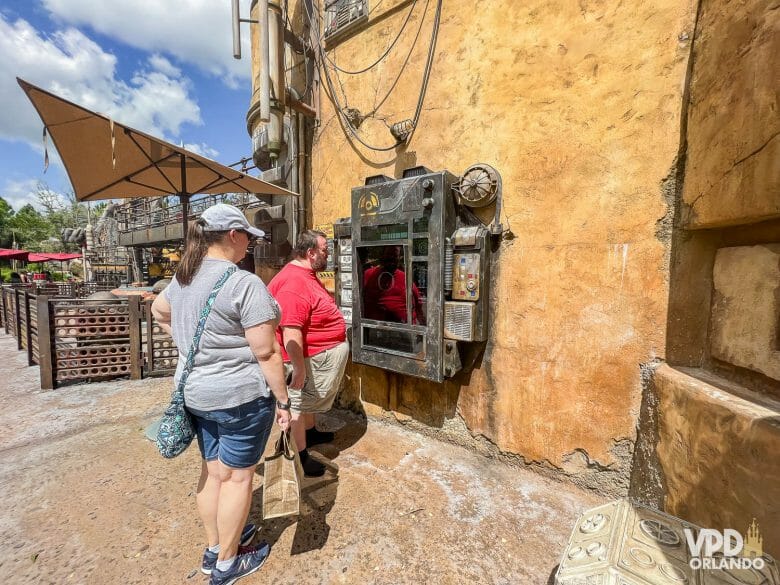 foto da tela onde os visitantes podem pegar missões no jogo batuu bounty hunters. há duas pessoas aguardando e um guarda-sol ao fundo.