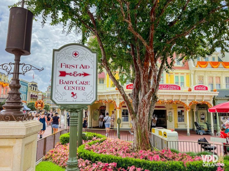 Foto da placa indicando o caminho do First Aid e do Baby Care Center no Magic Kingdom, áreas importantes pra conhecer quando se viaja com crianças. Ao fundo dá pra ver o Casey's Corner e a Main Street.