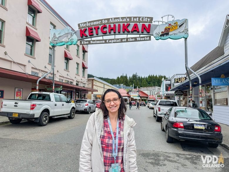 Renata na rua principal do porto de Ketchikan com placa ao fundo com o nome da cidade.